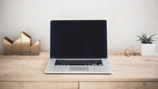 木製デスクの上に置かれた画面がオフのノートパソコンと観葉植物の写真