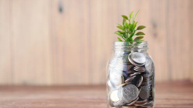 木製の背景の前に置かれたガラス瓶いっぱいのコインと、そこから伸びる緑の植物。お金を育てる・貯める・使うイメージ写真