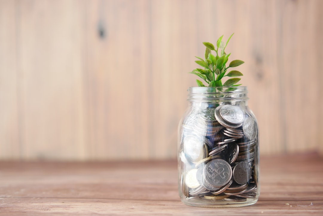 木製の背景の前に置かれたガラス瓶いっぱいのコインと、そこから伸びる緑の植物。お金を育てる・貯める・使うイメージ写真