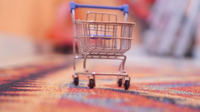カラフルな織り柄のラグの上に置かれた小さなショッピングカートのミニチュア。複数店舗での買い回りをイメージした写真